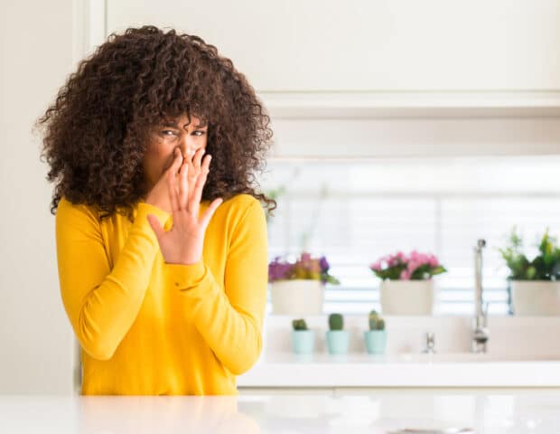 Young woman in a yellow top holding her nose in a kitchen, reacting to a bad smell.