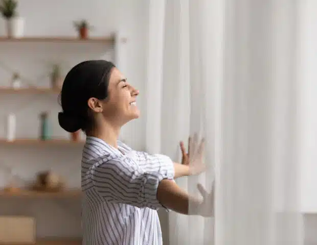 Excited young indian lady meet first morning at new flat house part curtains enjoy being homeowner. Happy hindu female open drapes on window breath fresh air close eyes in delight